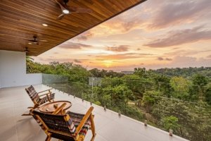 Modern house balcony with ocean view, sunset, and lush greenery.