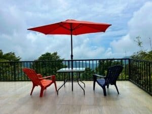 Balcony with red and blue chairs, table, and umbrella overlooking lush landscape.