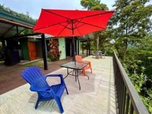 Outdoor balcony with colorful chairs, table, and red umbrella overlooking lush greenery.