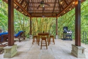 Covered outdoor porch with dining table, lounge chairs, and grill in lush jungle setting.