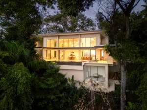 Modern tropical home with large glass windows surrounded by lush greenery.