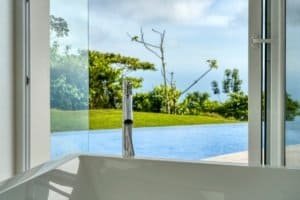 Modern bathroom with ocean view, glass doors, and lush greenery.