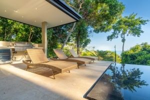 Modern outdoor terrace with lounge chairs and infinity pool overlooking lush greenery and ocean.