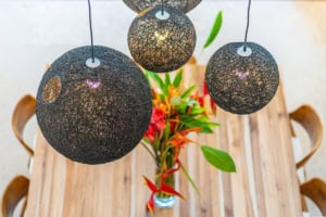 Three black woven pendant lights hanging above a wooden dining table with tropical plants.