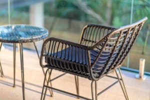 Wicker chair and glass table on balcony with lush greenery view.