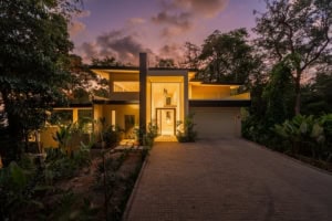 Modern tropical house with large windows and lush garden at sunset.