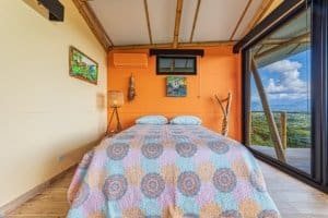 Bedroom with large window overlooking lush mountain landscape and blue sky.