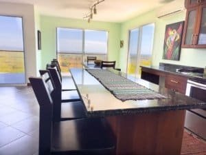 Bright dining room with panoramic ocean and mountain views from large windows.
