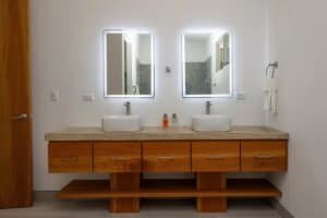 Contemporary bathroom with dual sinks, illuminated mirrors, and wooden vanity.