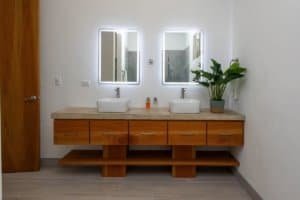 Contemporary bathroom with dual sinks, illuminated mirrors, and wooden vanity.