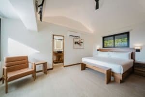 Modern bedroom with king bed, wooden furniture, and large window with jungle view.