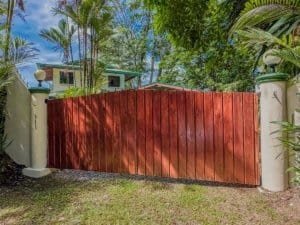 Red wooden gate with lush tropical trees and house in background.