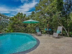 Outdoor pool with lounge chairs and umbrella in lush tropical setting.