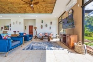 Bright living room with blue sofas, large TV, and ocean view through glass doors.