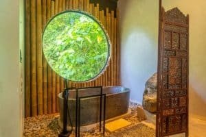 Japanese style home bathtub with lush jungle view through round window.