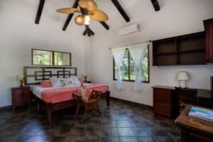 Interior of a Bali style bedroom with pink bedding, ceiling fan, and large windows.