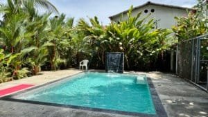 Rectangle swimming pool with tiled edge surrounded by tropical plants and a white plastic chair.