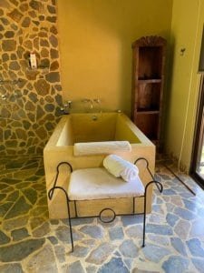 Stone-tiled bathroom with sunlit bathtub and towel on stand, wooden cabinet nearby.