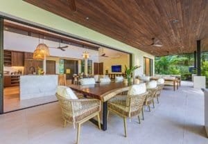 Ocean view villa patio with wooden ceiling, wicker chairs around a long dining table and open living.