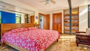 Bedroom with red floral bedspread, wooden furniture, stone floor, shelves, and ceiling fan in a suny.
