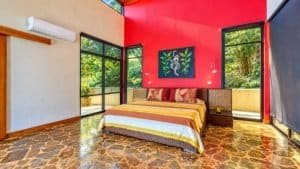 Bright bedroom with red accent wall, stone floor, and large windows showing lush tropical views.