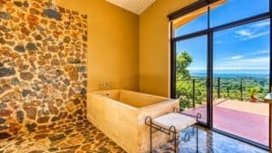 Stone-tiled bathroom with large window overlooking lush valley and ocean view.