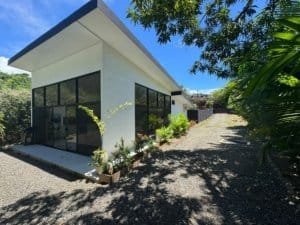 Modern single-story villa with large glass windows, white walls, gravel driveway, and tropical trees.