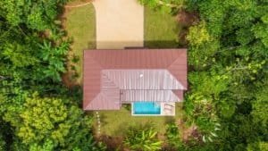Aerial view of a brown-roofed house with a blue pool, surrounded by dense tropical trees.