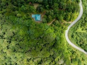 Aerial view of a house with turquoise roof surrounded by dense forest and a winding road.