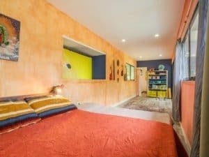 Bright bedroom with orange walls, colorful artwork, and large window.