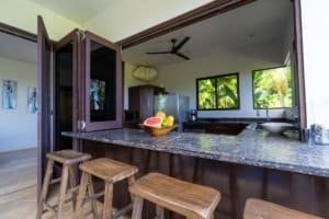 Modern kitchen with ocean view, tropical greenery, and bar stools.