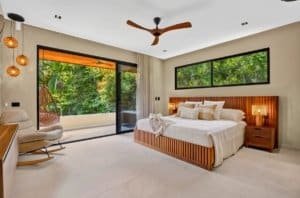 Luxurious king-size bedroom with wooden headboard, large windows, and tropical greenery outside.