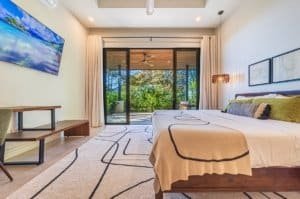 Bright hotel room with king bed, wall-mounted TV, and sliding glass doors to lush garden.
