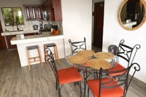 Dining area with glass-top table and four chairs near kitchen, coastal home interior.