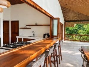 Open-air kitchen with wood counter, bar stools, and lush green backdrop in Costa Rica.
