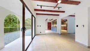 Bright open living area with large glass doors to outdoors, white walls, exposed wooden beams, and a.