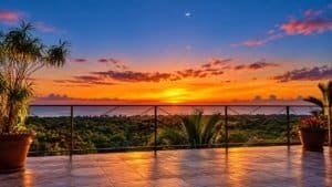 Sunset over tropical hillside ocean view from terrace with plants and railing.