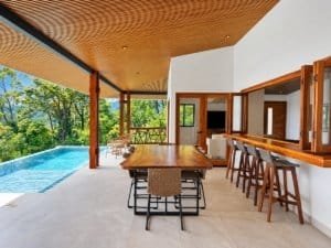 Open-air dining area with wooden table and bar stools by a pool and lush jungle in Dominical, Costa.