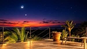 Panoramic terrace with sunset over horizon, palm plants and starry night sky.