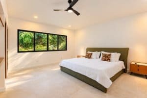 Bright master bedroom with large window overlooking lush greenery and a ceiling fan.