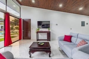 Spacious living room with white marble floors, grey sofa, red cushions, and large glass doors.