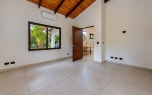 Bright empty living space with large window, wooden ceiling beams and tiled floor.