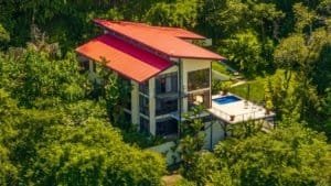 Modern hillside villa with red roof amid lush tropical trees and a private pool.
