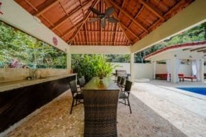 Outdoor covered dining area with pool and lush tropical surroundings.
