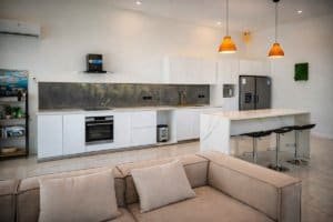Open plan kitchen with white cabinets, marble island, bar stools and pendant lights.