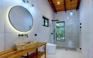 Bright bathroom with round mirror, stone vessel sink, wooden vanity, and glass shower in tropical 4.