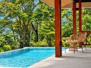 Ocean view infinity pool with tiled terrace and wooden pillars on lush green hillside.