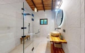 Modern tropical bathroom with glass shower, circular backlit mirror, wooden vanity and skylight.
