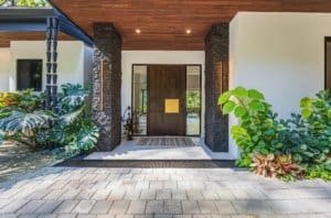 Modern villa entrance with stone columns, wooden door, and tropical greenery.