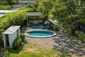 Backyard with kidney-shaped pool, surrounding gravel yard, shaded by trees and a modern glass-walled.